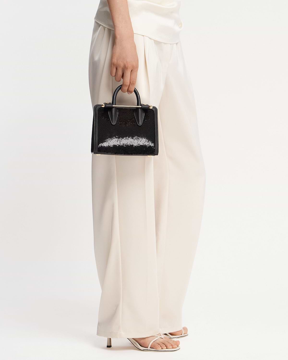 Hand holding black sequinned nano tote with top handles against white trousers.