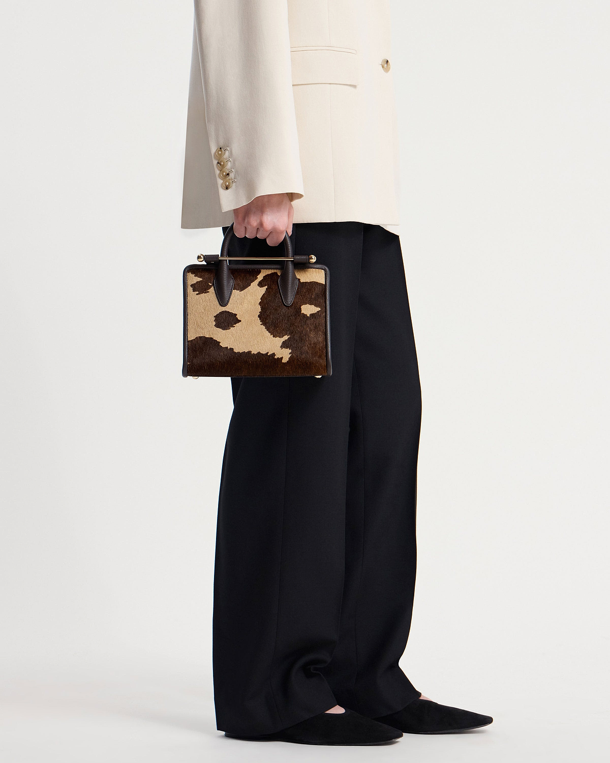 Model hand holding sand and espresso spot print mini tote, cream blazer sleeve visible.