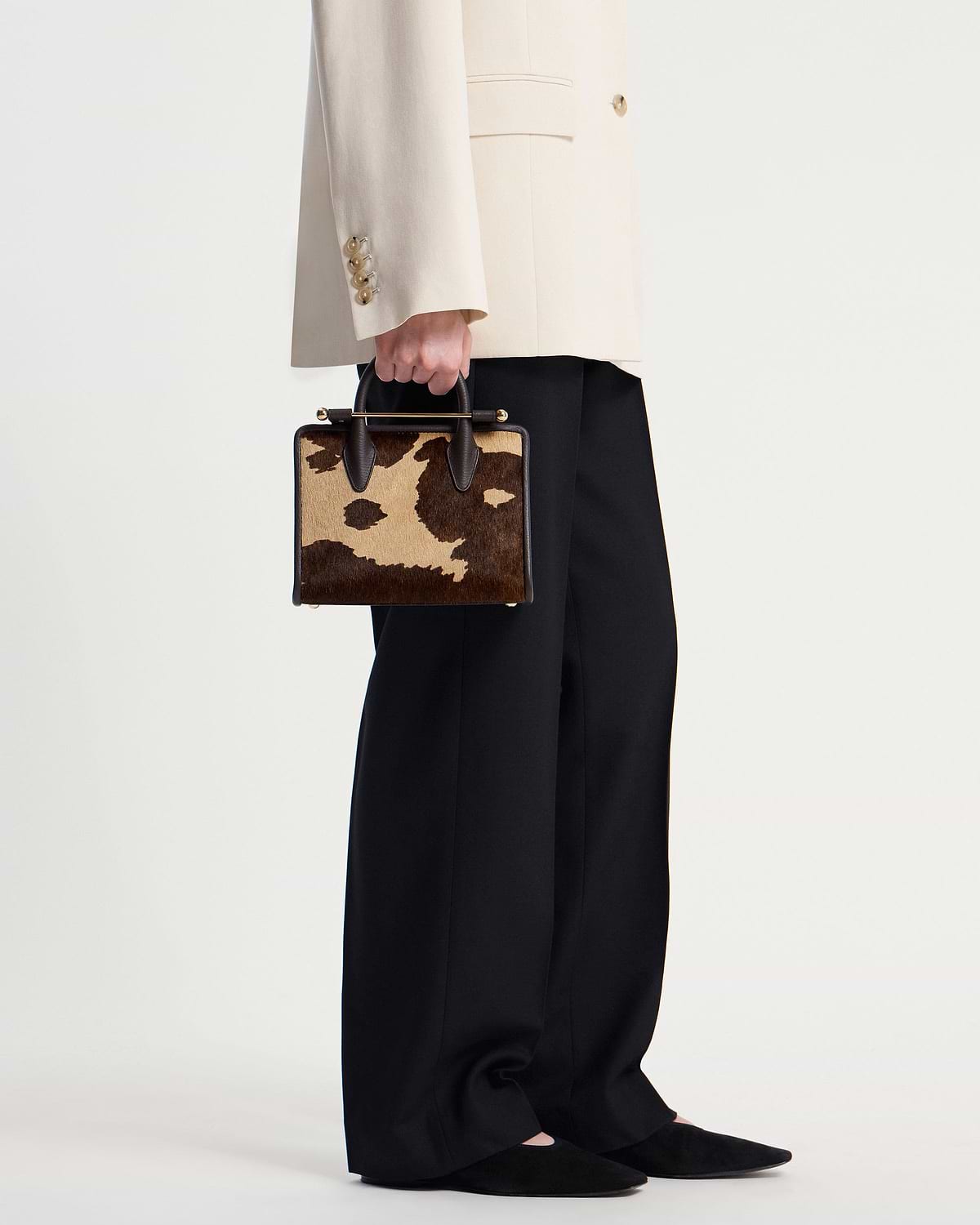 Model hand holding sand and espresso spot print mini tote, cream blazer sleeve visible.