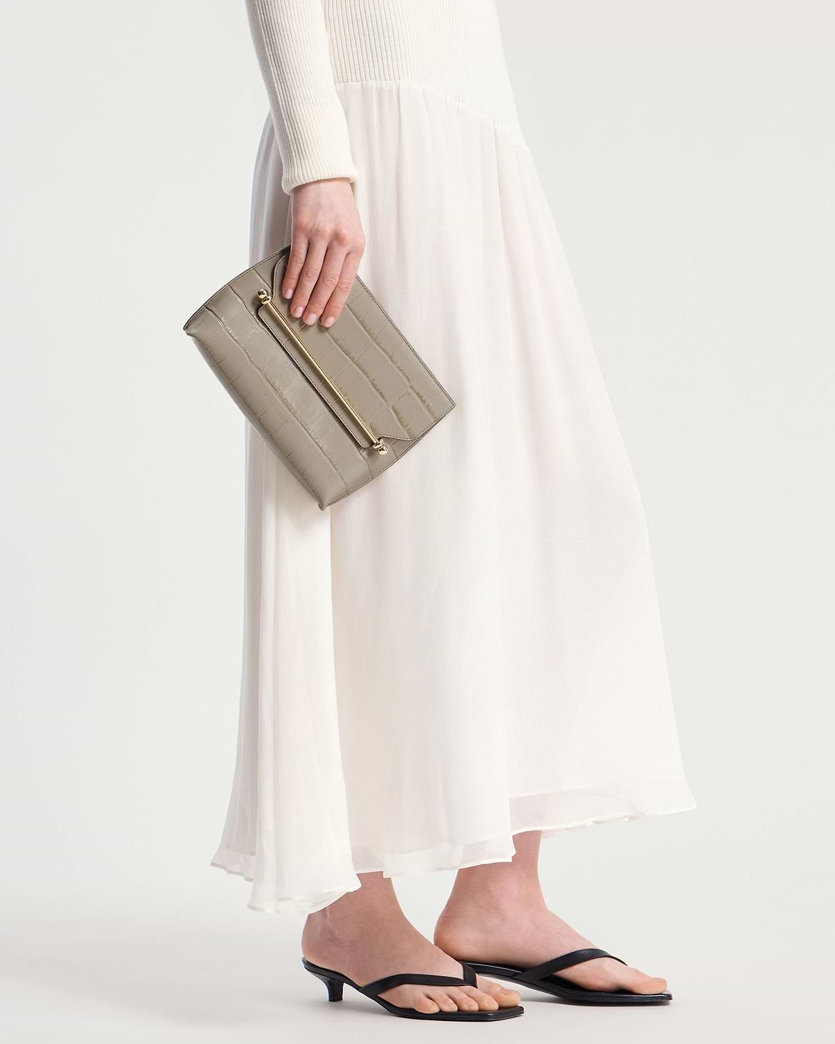 Hand holding croc-embossed clutch with gold bar, model in white dress.