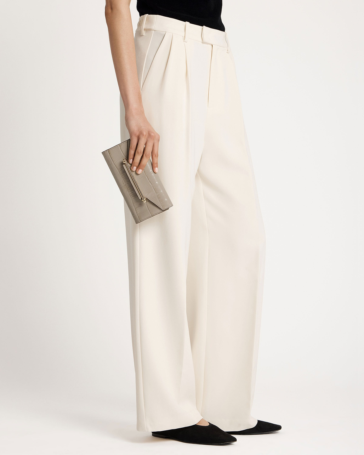 Model holding a croc-embossed grey wallet at hip with cream wide-leg trousers