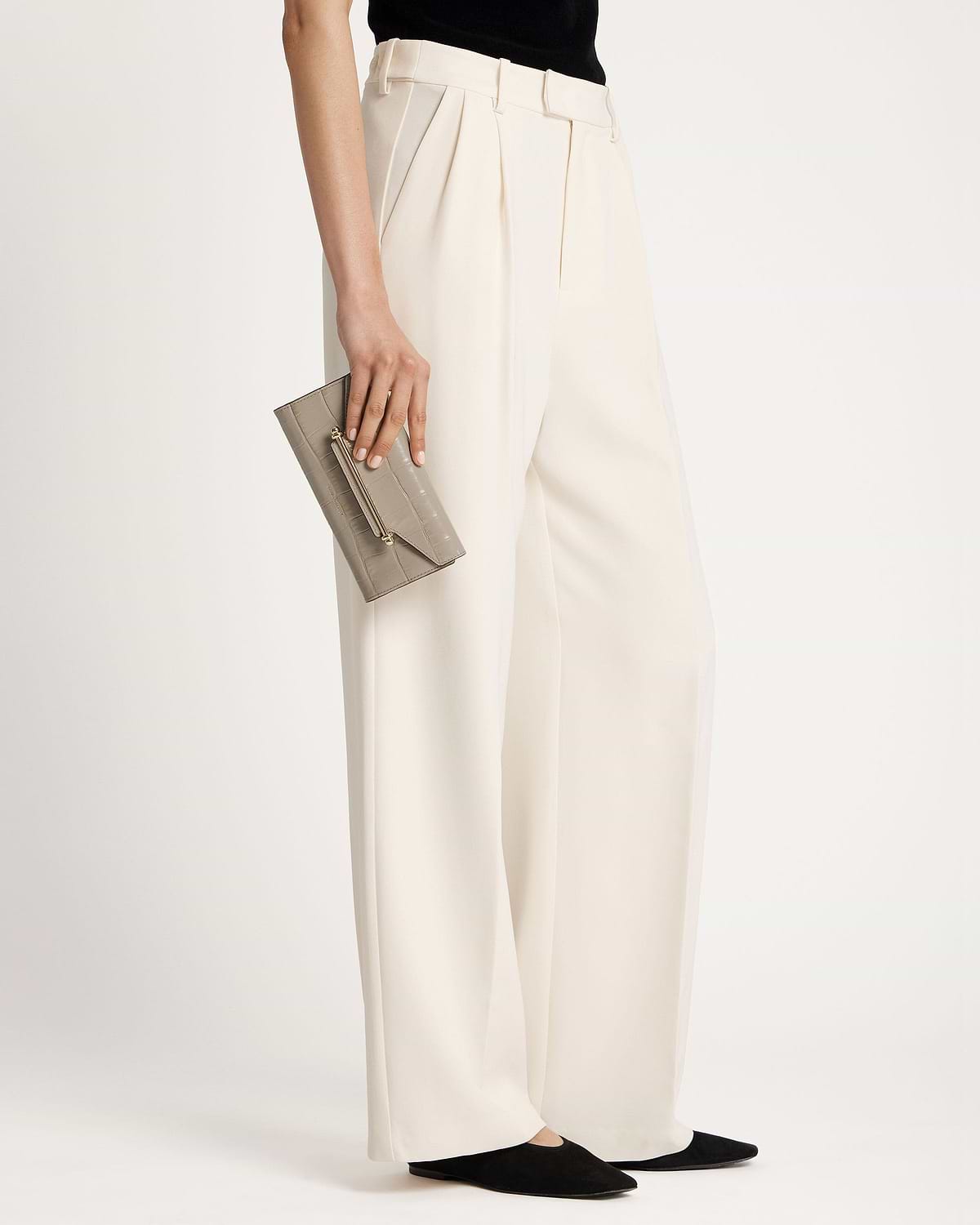 Model holding a croc-embossed grey wallet at hip with cream wide-leg trousers