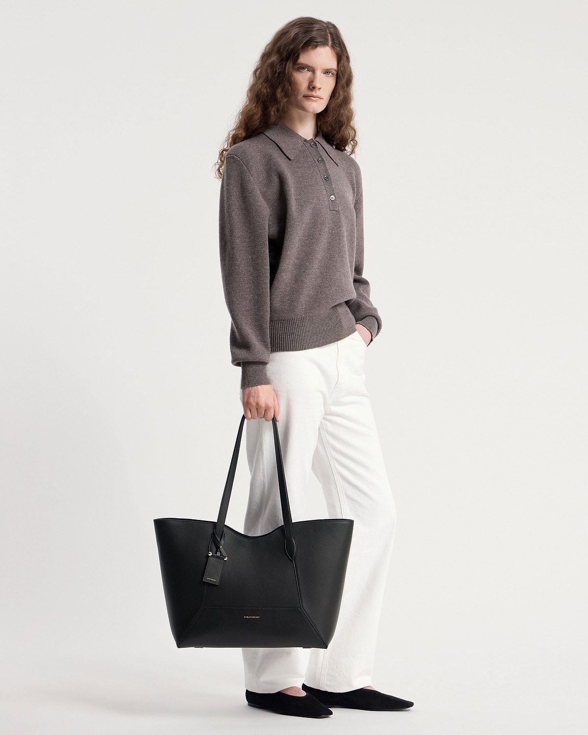Model holding black Strathberry Mosaic Cabas tote by handles, wearing white trousers.
