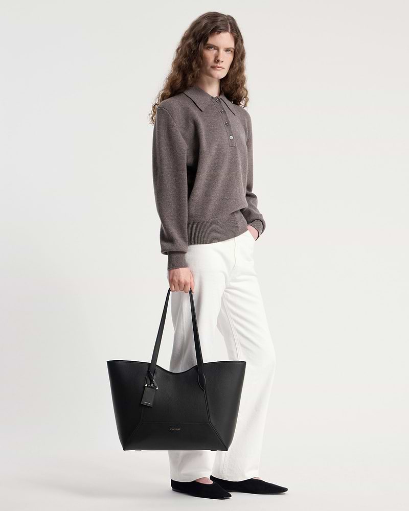 Model holding black Strathberry Mosaic Cabas tote by handles, wearing white trousers.