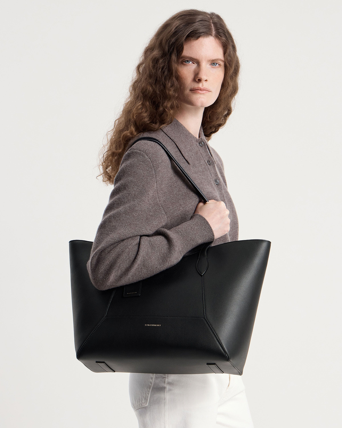 Model wearing brown jumper holding black Strathberry tote bag over shoulder.