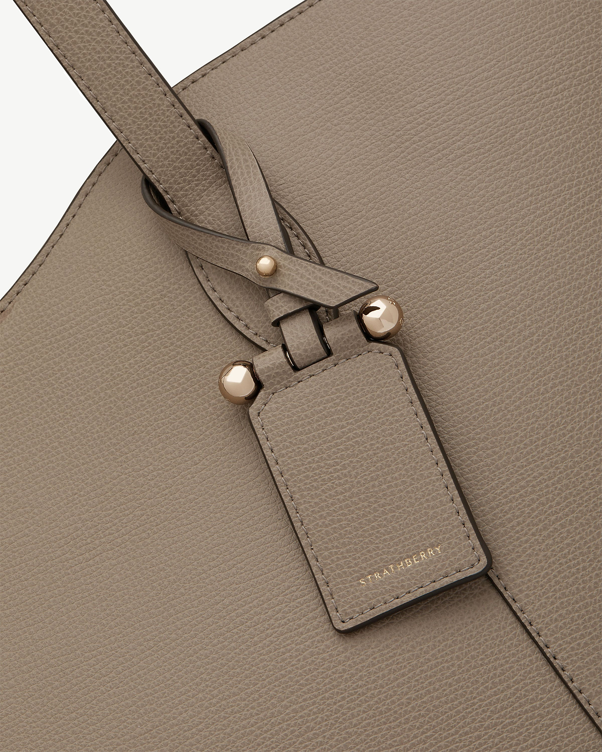 Taupe pebbled leather strap with gold studs and Strathberry luggage tag.