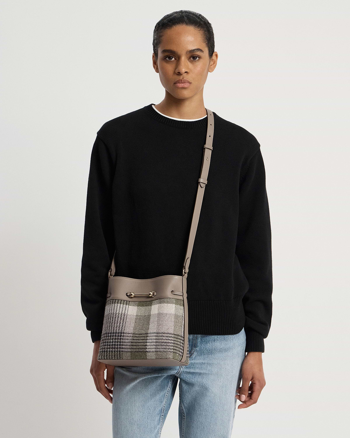Model wearing taupe leather-strapped tartan bucket bag with gold bar detail across body