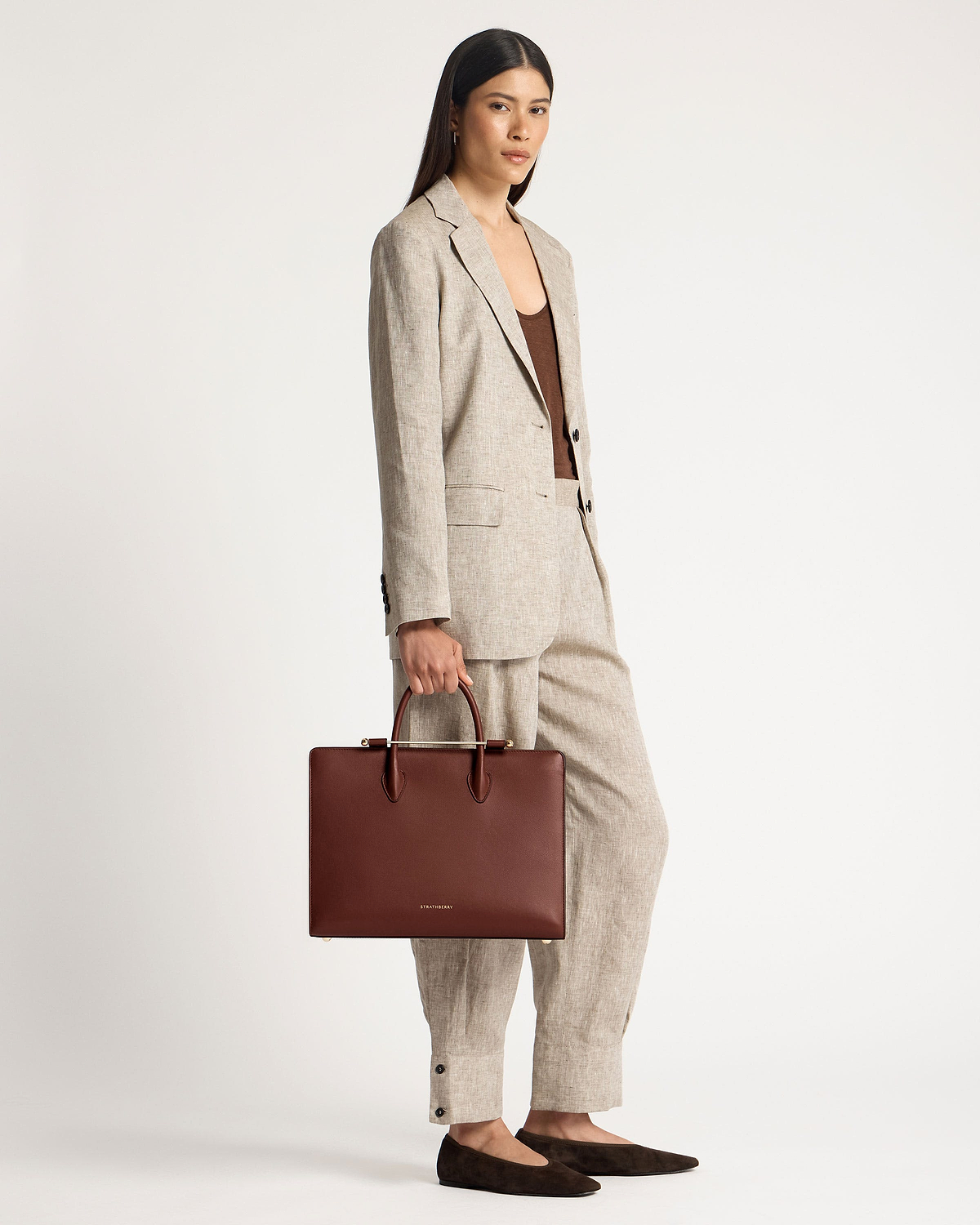Model holding walnut tote bag with Strathberry logo, wearing a linen suit