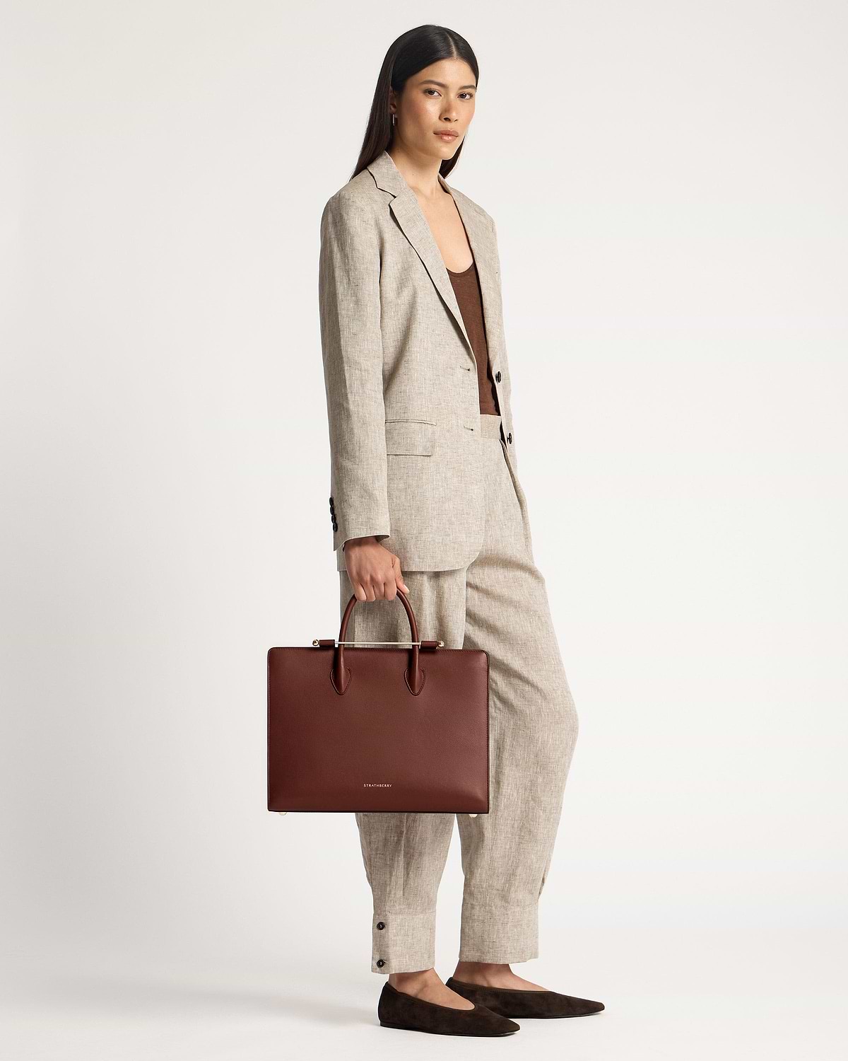 Model holding walnut tote bag with Strathberry logo, wearing a linen suit