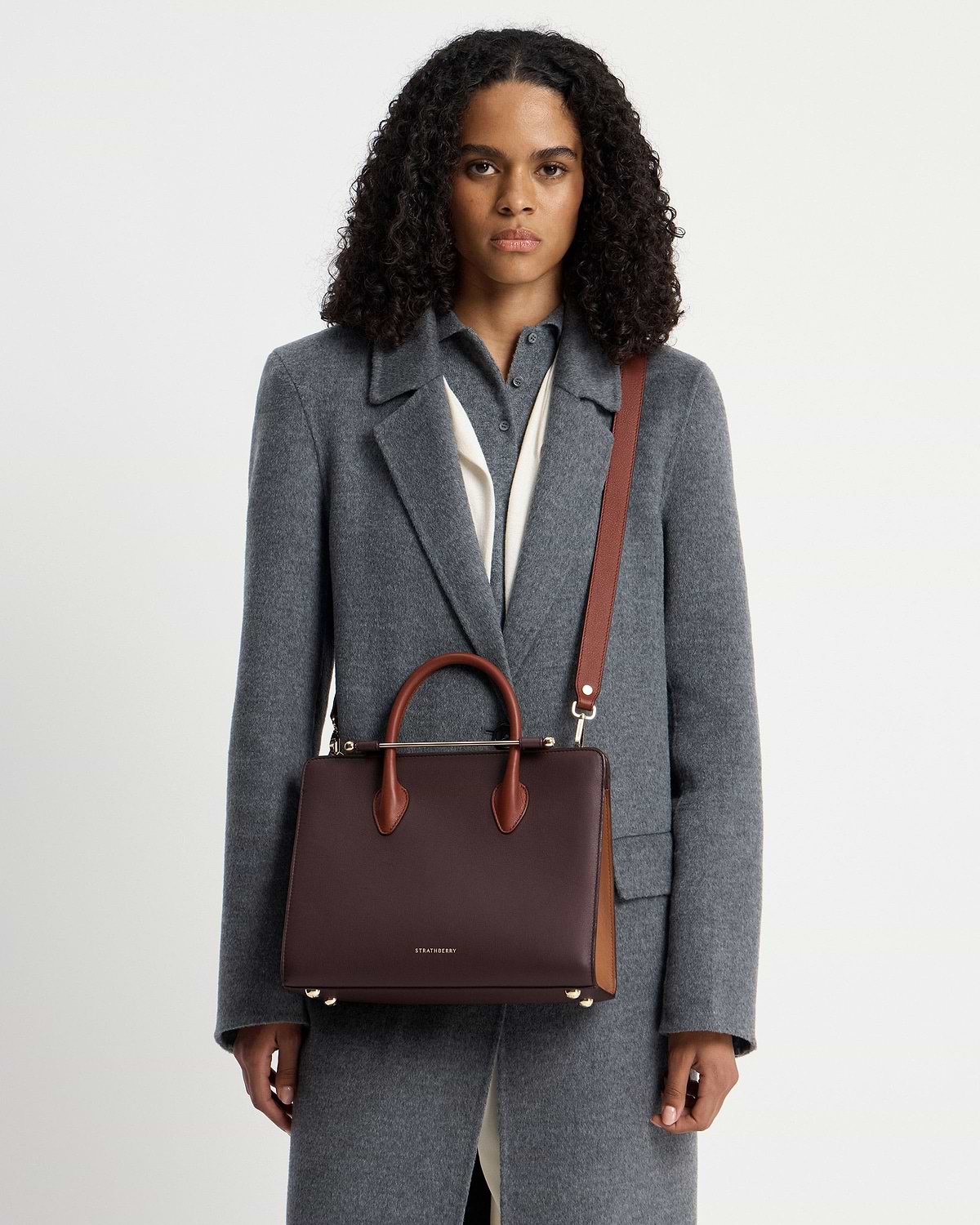 Model wearing grey coat holding Strathberry midi tote in burgundy, walnut and chestnut