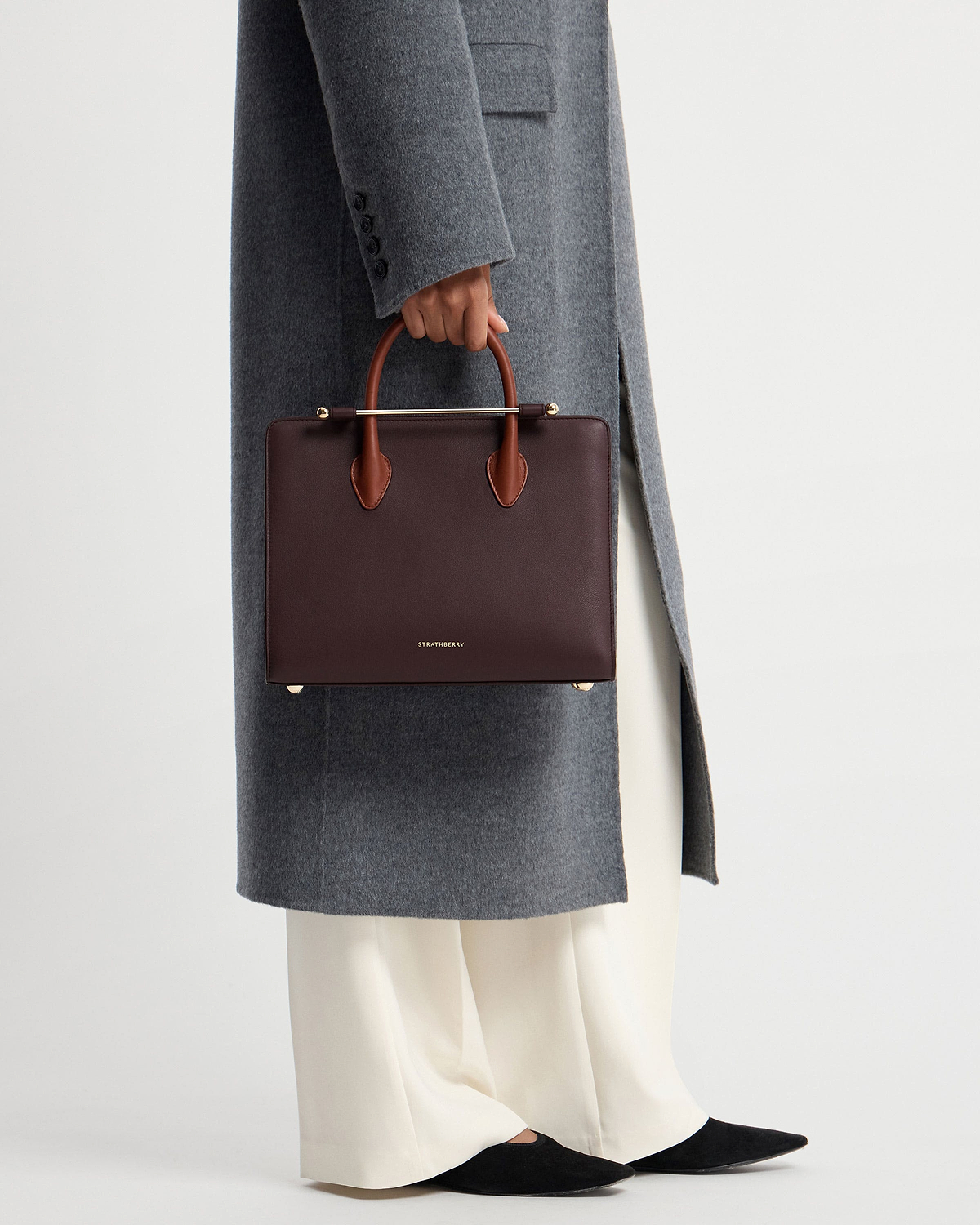 Model waist down holding burgundy Strathberry midi tote with chestnut handles and gold bar.