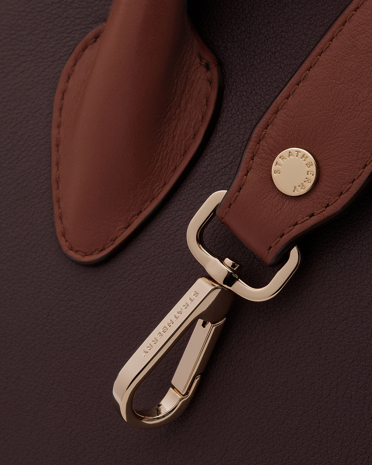 Chestnut leather strap on burgundy background with gold hardware, Strathberry button.