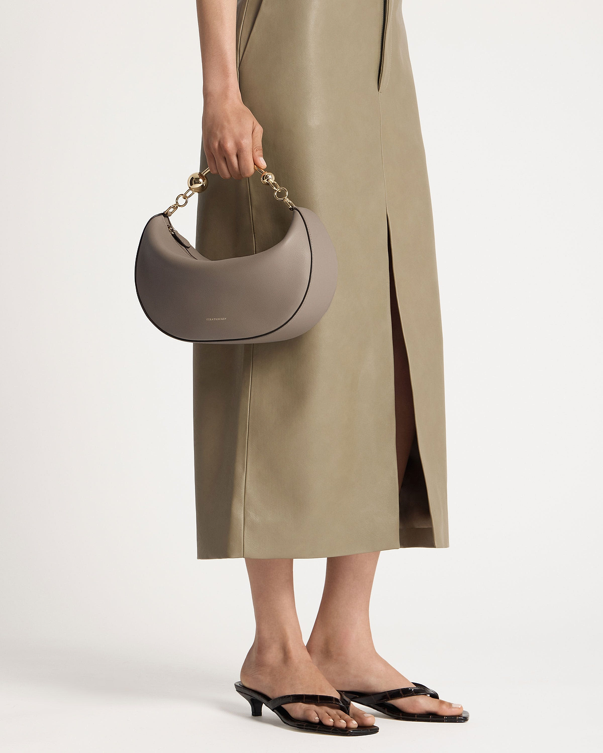 Model holding taupe crescent-shaped handbag with gold chain handle and olive midi skirt.