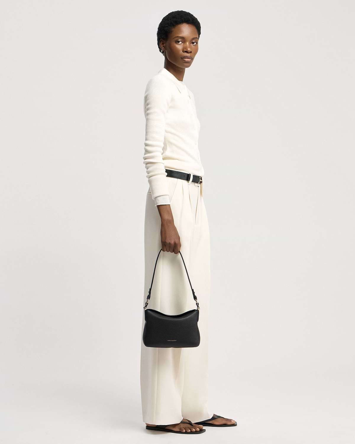 Model holding black Sway shoulder bag by Strathberry, wearing white top and cream trousers.