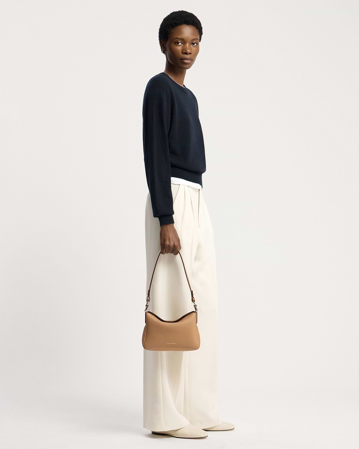 Model holding a caramel Strathberry Sway shoulder bag with top handle and visible logo.