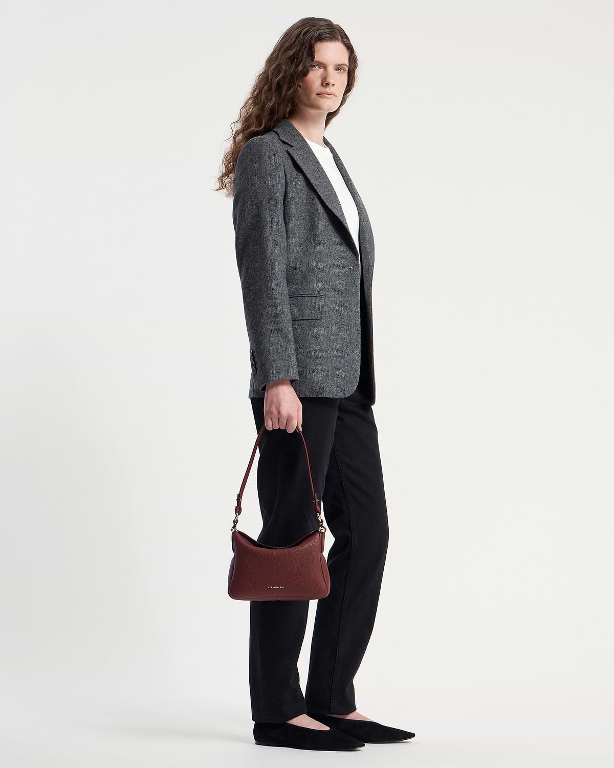 Model holding oxblood Sway shoulder bag by Strathberry while wearing grey blazer.