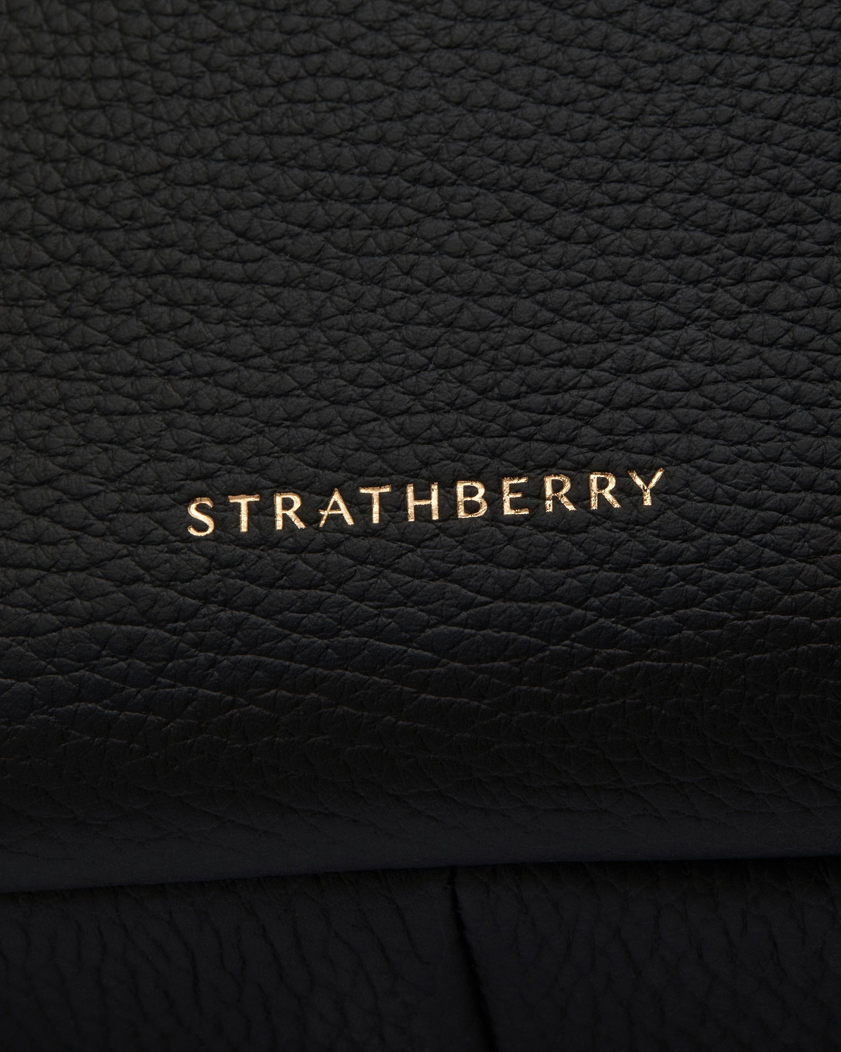 Close-up of black pebbled leather with gold Strathberry logo embossed near the bottom.