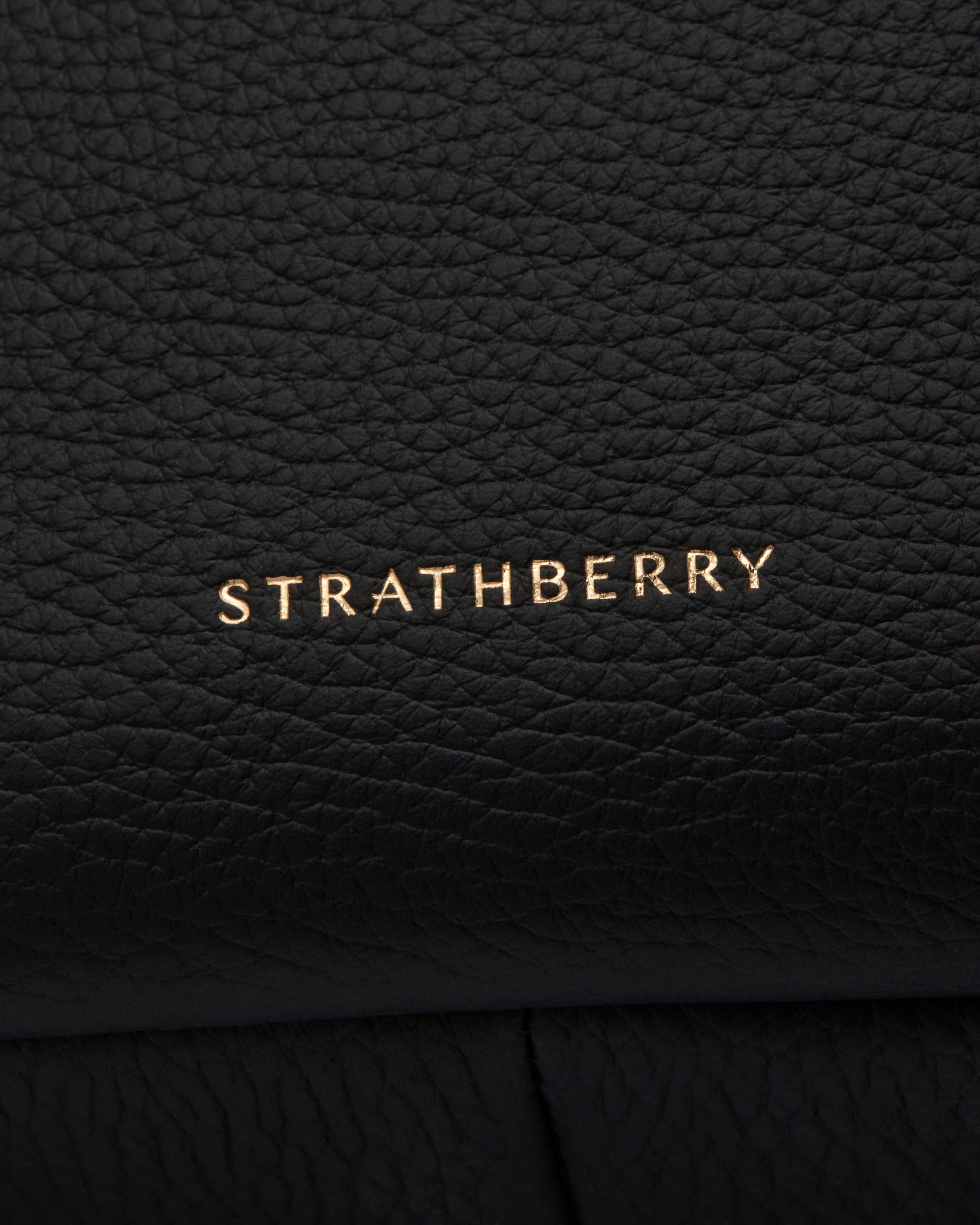 Close-up of black pebbled leather with gold Strathberry logo embossed near the bottom.