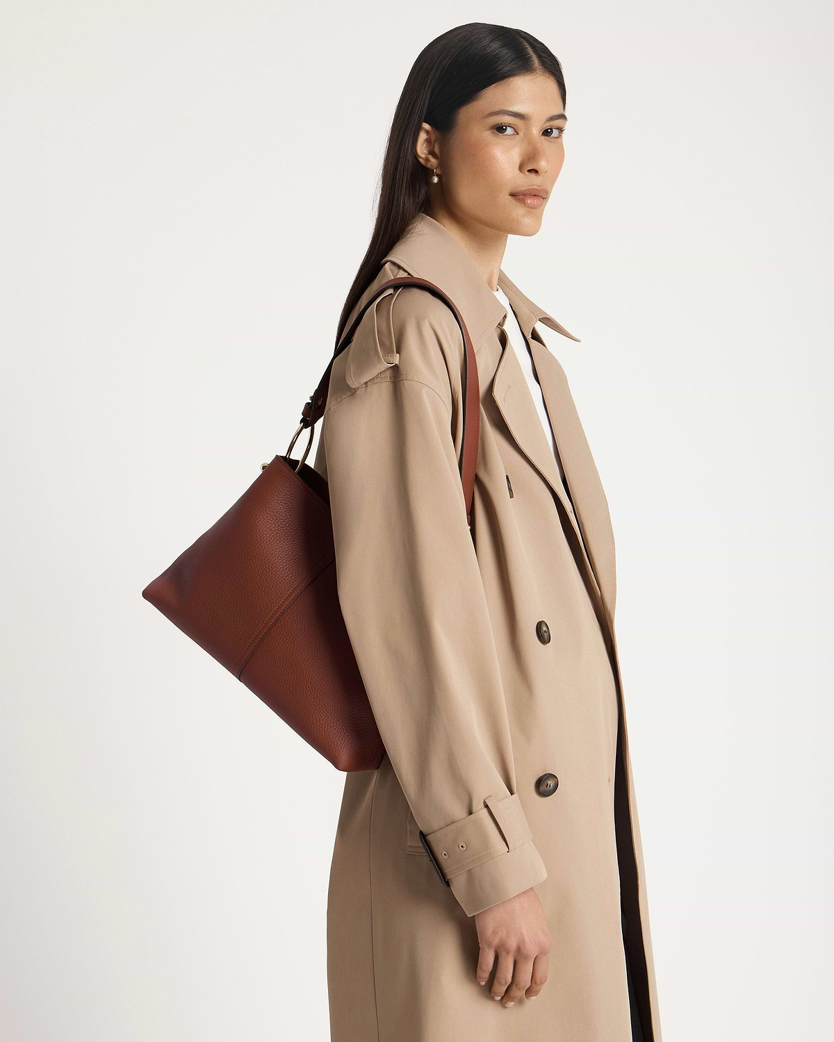 Model wearing beige trench coat carrying walnut brown leather hobo bag on shoulder