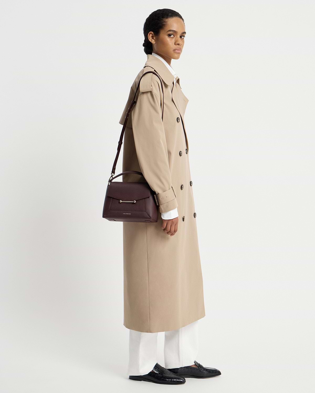 Model in beige trench coat carrying burgundy Strathberry Mosaic bag on shoulder