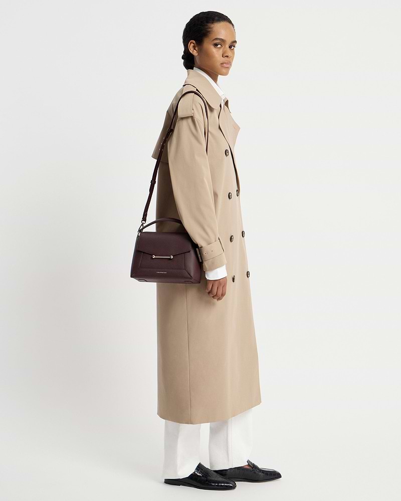 Model in beige trench coat carrying burgundy Strathberry Mosaic bag on shoulder