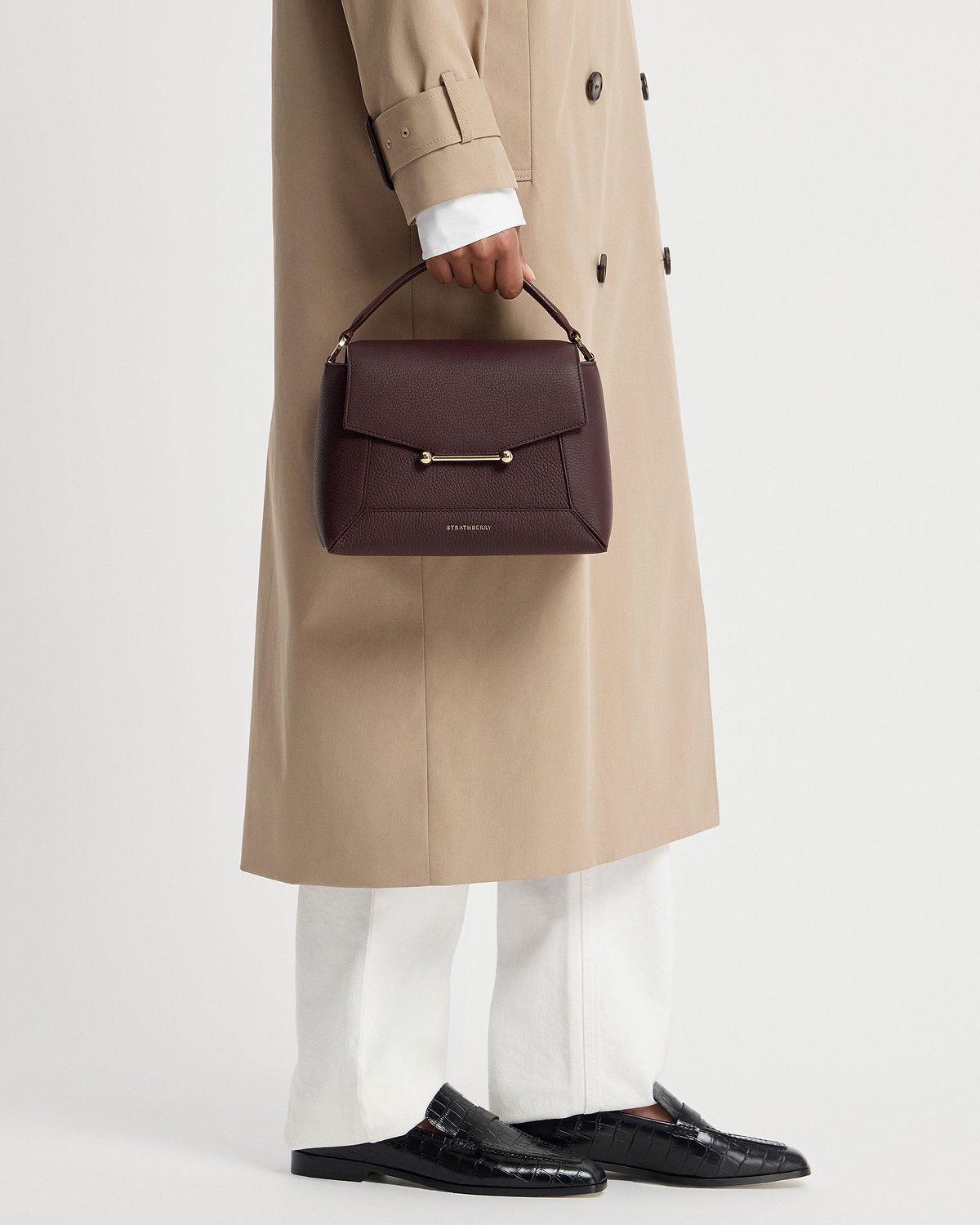 Model holding burgundy Strathberry Mosaic top-handle bag with gold bar detail.
