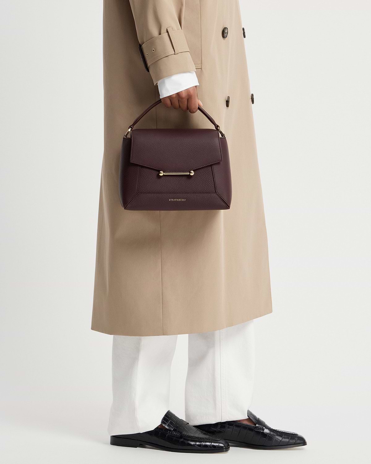Model holding burgundy Strathberry Mosaic top-handle bag with gold bar detail.