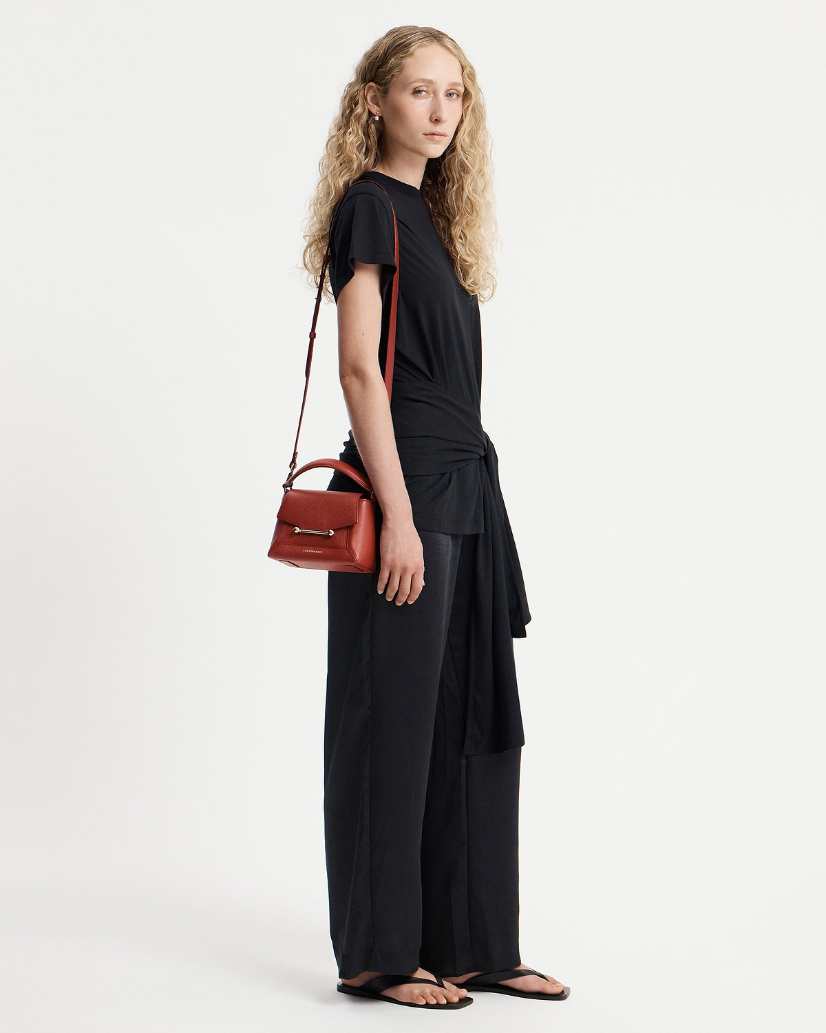 Model in black outfit carrying small red top-handle bag on her shoulder.
