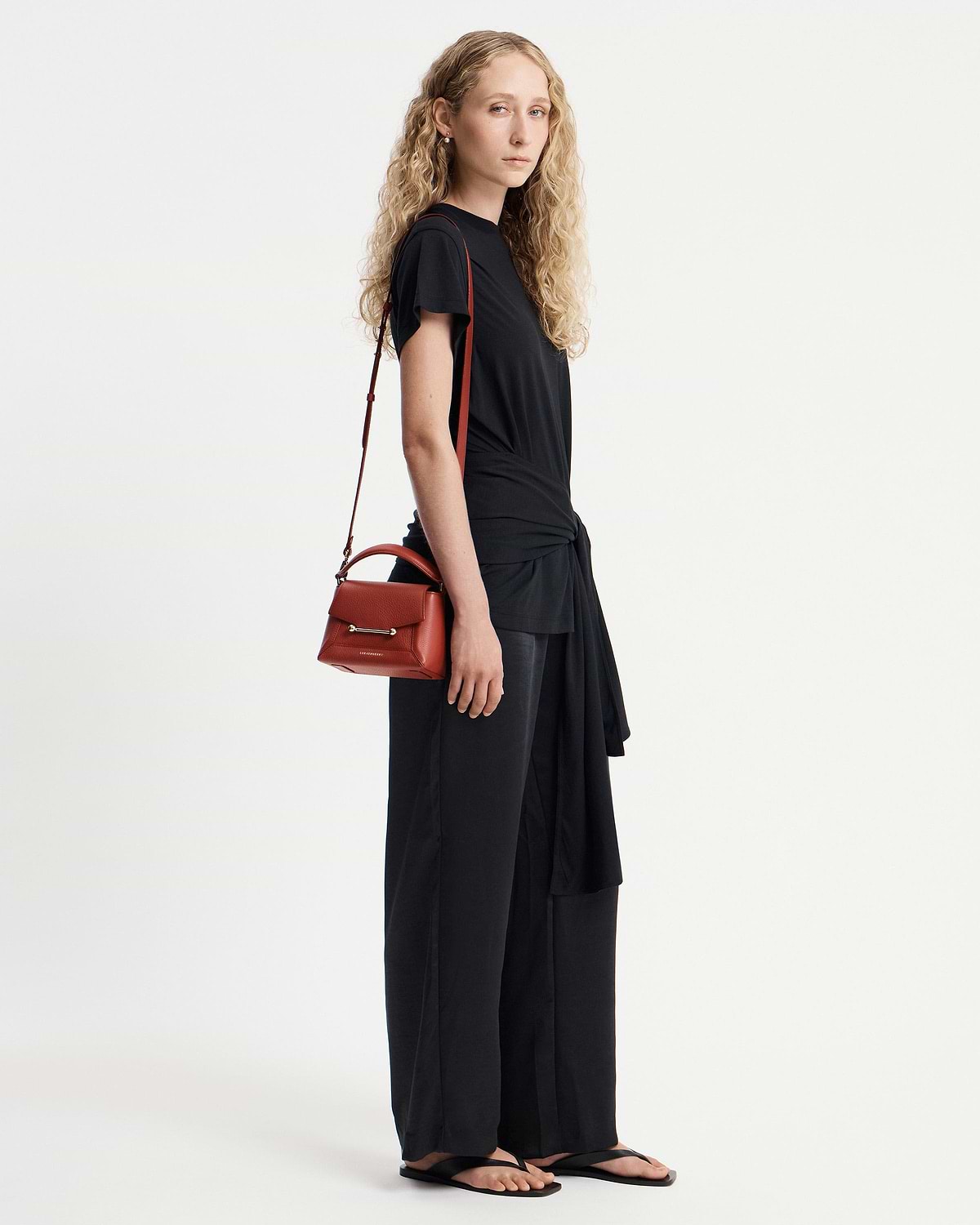 Model in black outfit carrying small red top-handle bag on her shoulder.