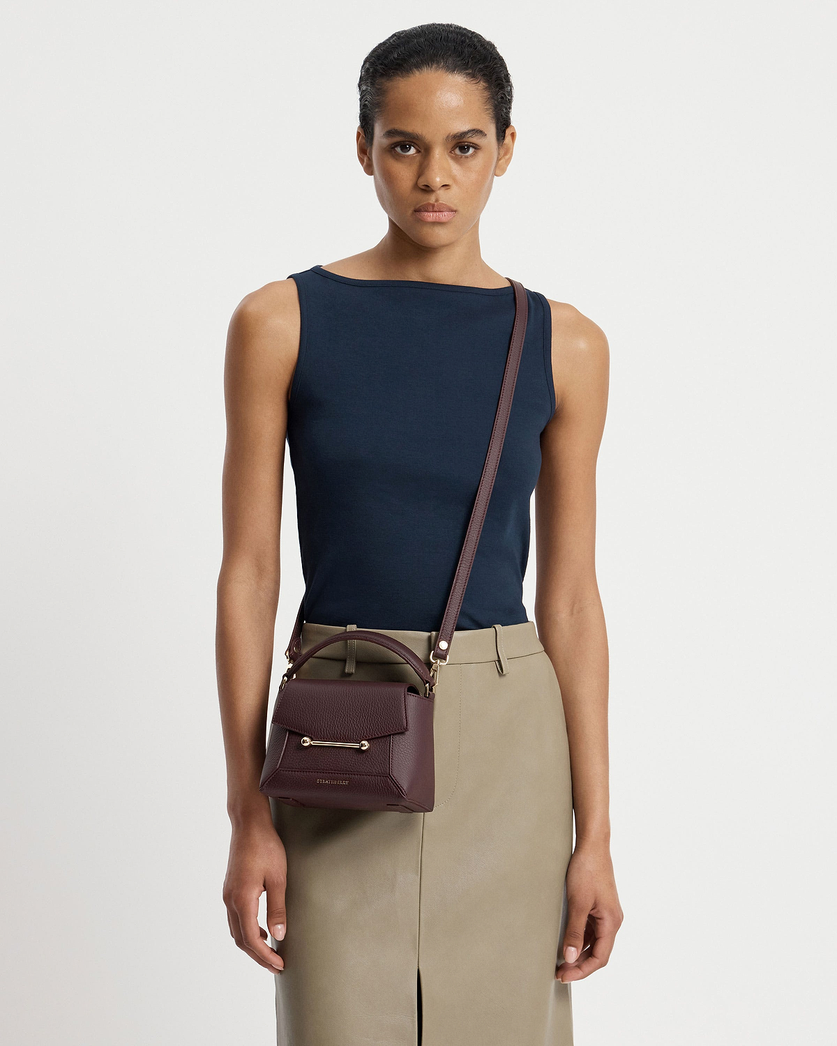 Model wearing navy top and beige skirt holds a burgundy Strathberry Mosaic Nano crossbody bag.