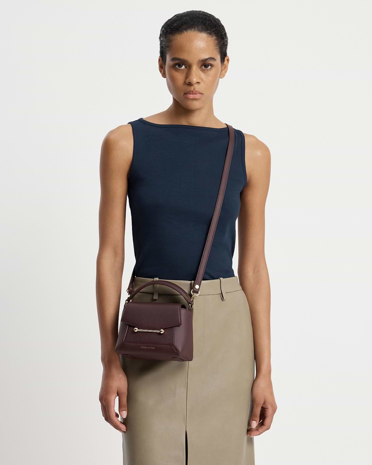 Model wearing navy top and beige skirt holds a burgundy Strathberry Mosaic Nano crossbody bag.