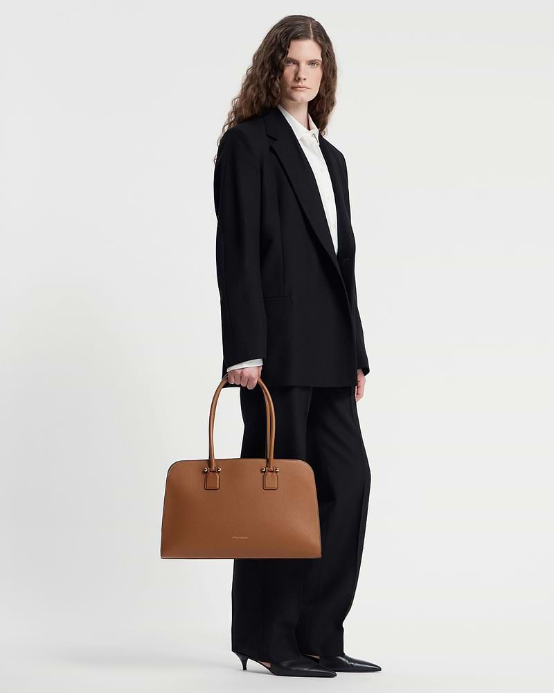 Model holding tan Georgia Maxi tote bag with Strathberry logo and double handles.