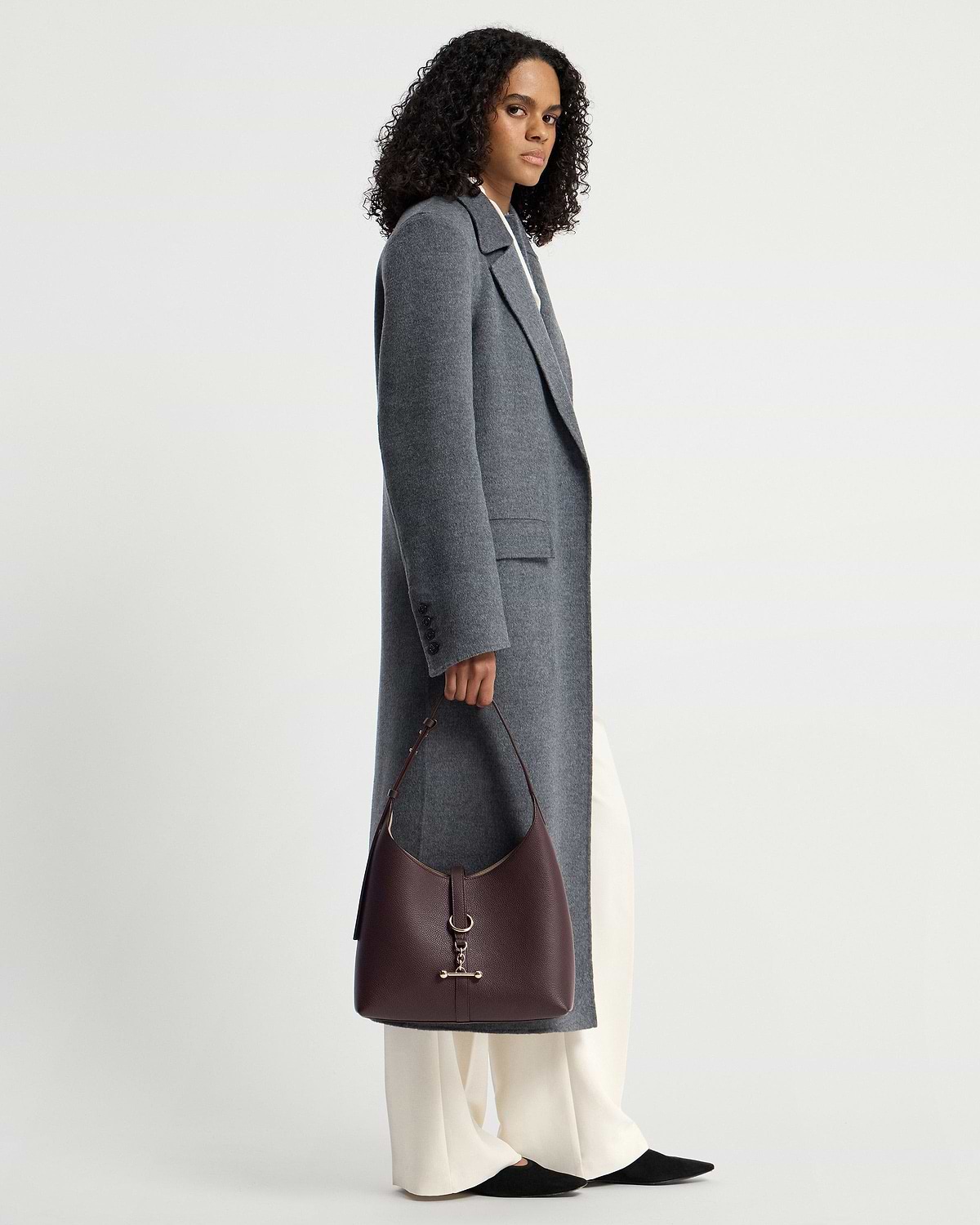 Model holding burgundy hobo bag with gold hardware, wearing grey coat and cream trousers.