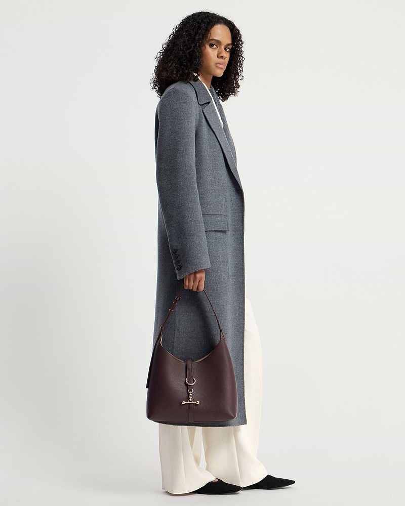 Model holding burgundy hobo bag with gold hardware, wearing grey coat and cream trousers.