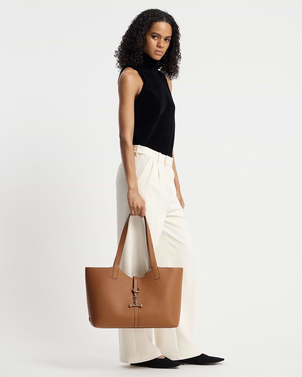 Model holding tan kite tote at her side, wearing black sleeveless top.