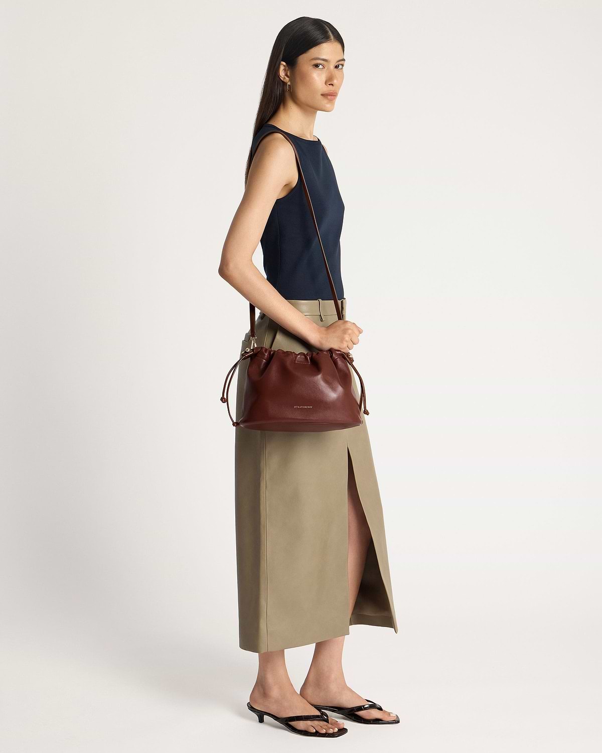 Woman wearing navy top and khaki skirt carrying walnut Strathberry Charlotte Midi drawstring bag.