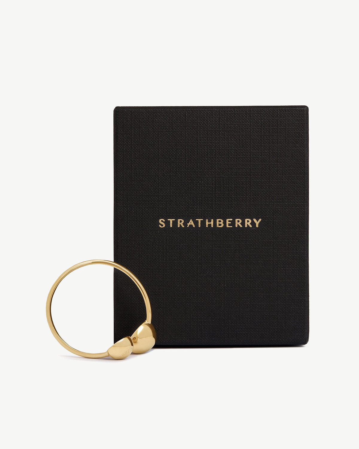 Light gold metal napkin ring beside black textured box with Strathberry logo.