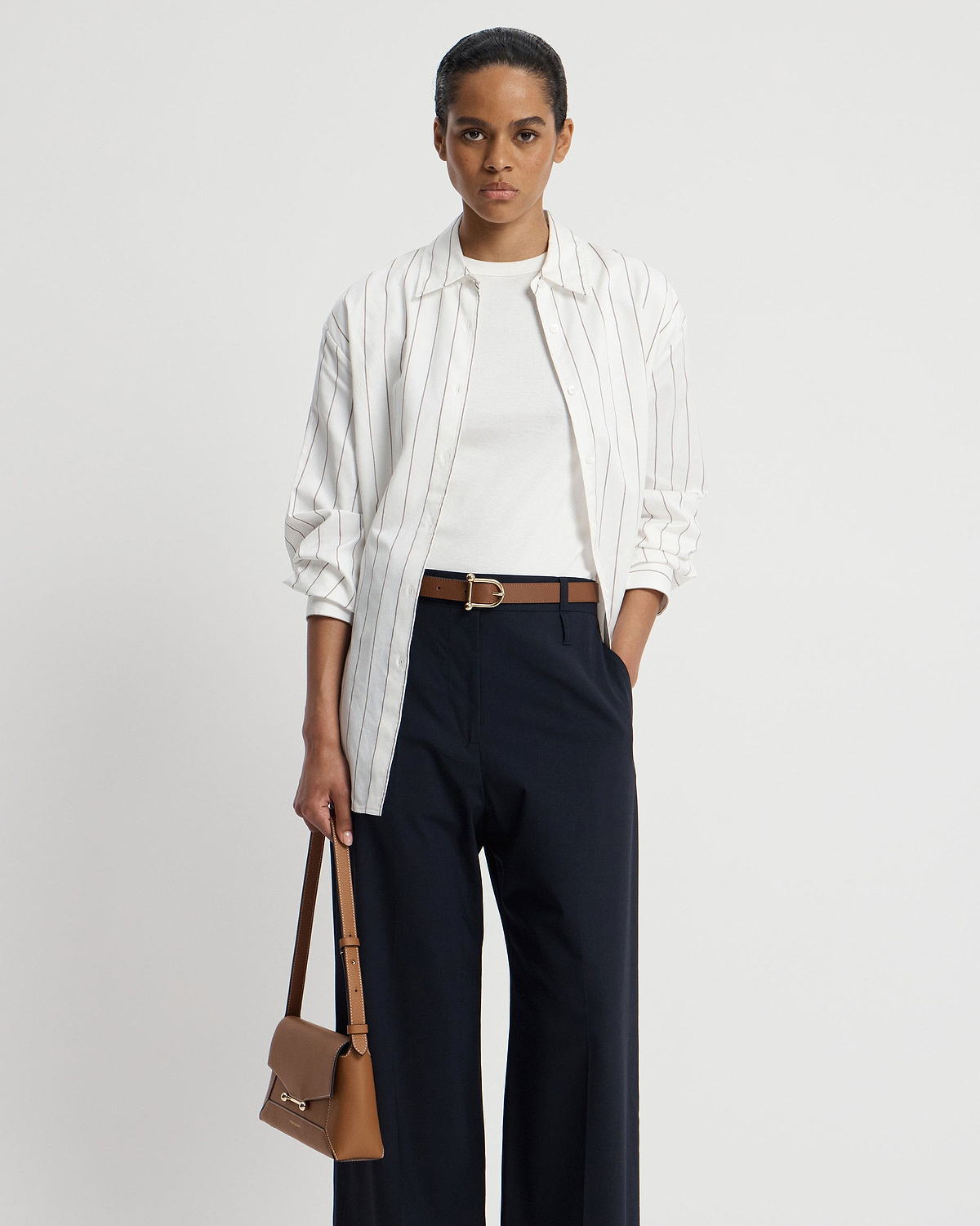 Model wearing hazelnut leather belt with gold buckle over navy trousers.