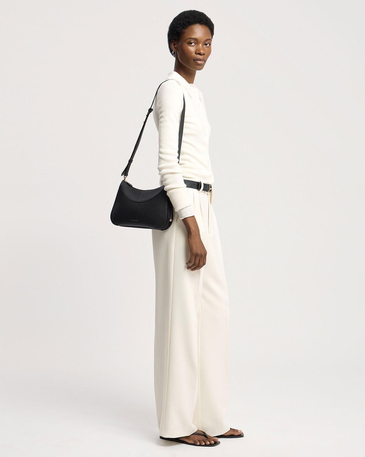 Model in white outfit carrying black Strathberry Nest shoulder bag on shoulder.