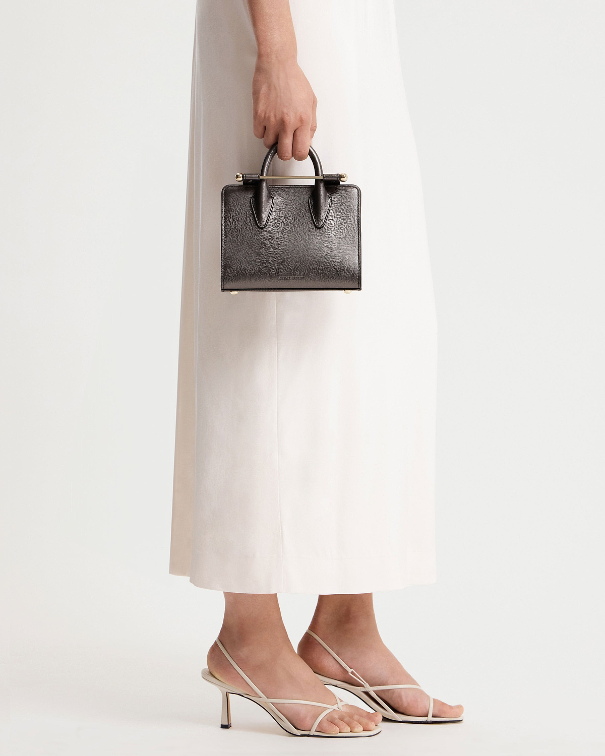 Hand holding metallic anthracite caviar Strathberry nano tote against white dress.