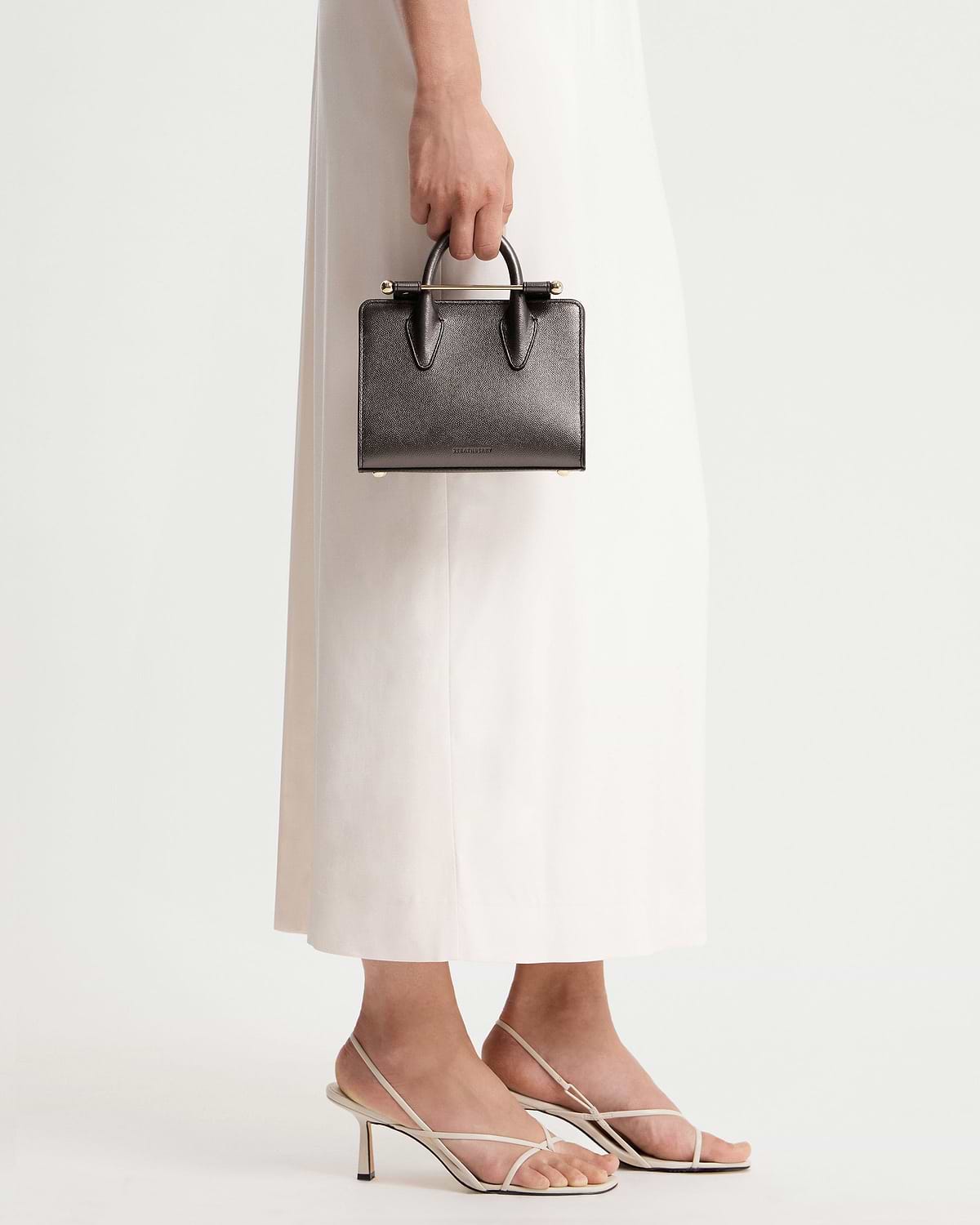 Hand holding metallic anthracite caviar Strathberry nano tote against white dress.