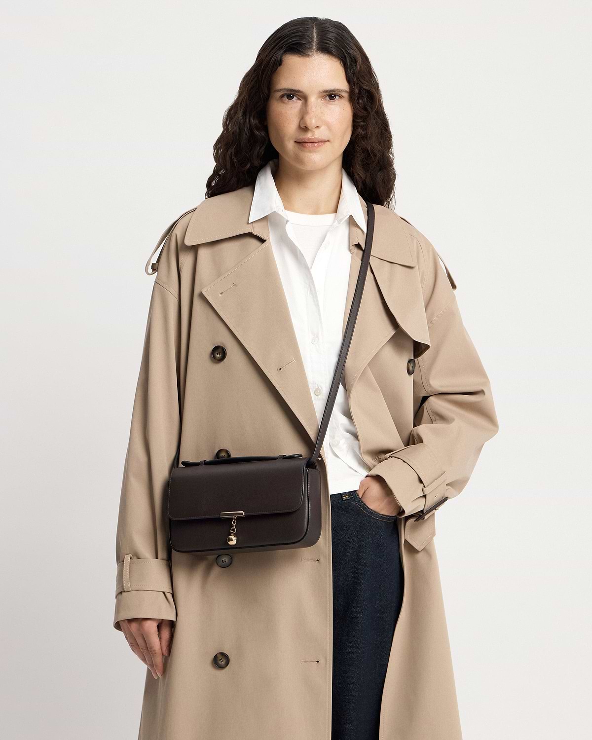 Model facing camera wearing beige trench coat and espresso clutch bag worn crossbody.