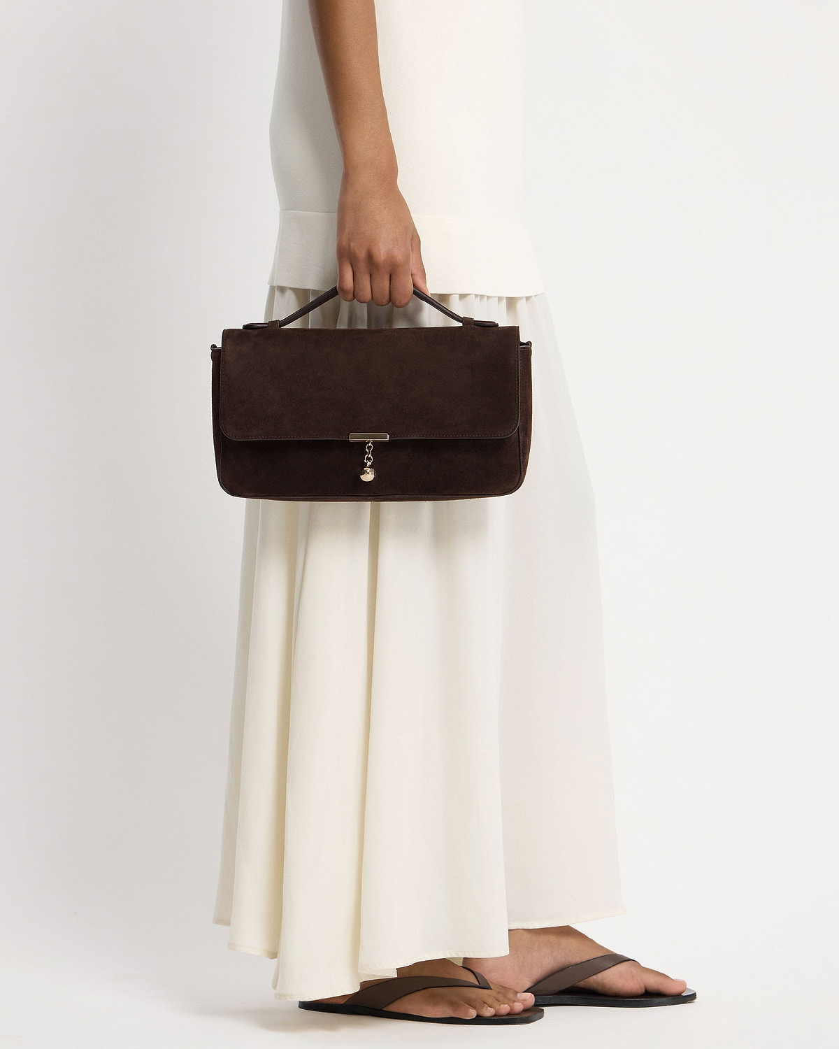 Hand holding espresso suede top-handle bag with gold charm, cream dress visible.