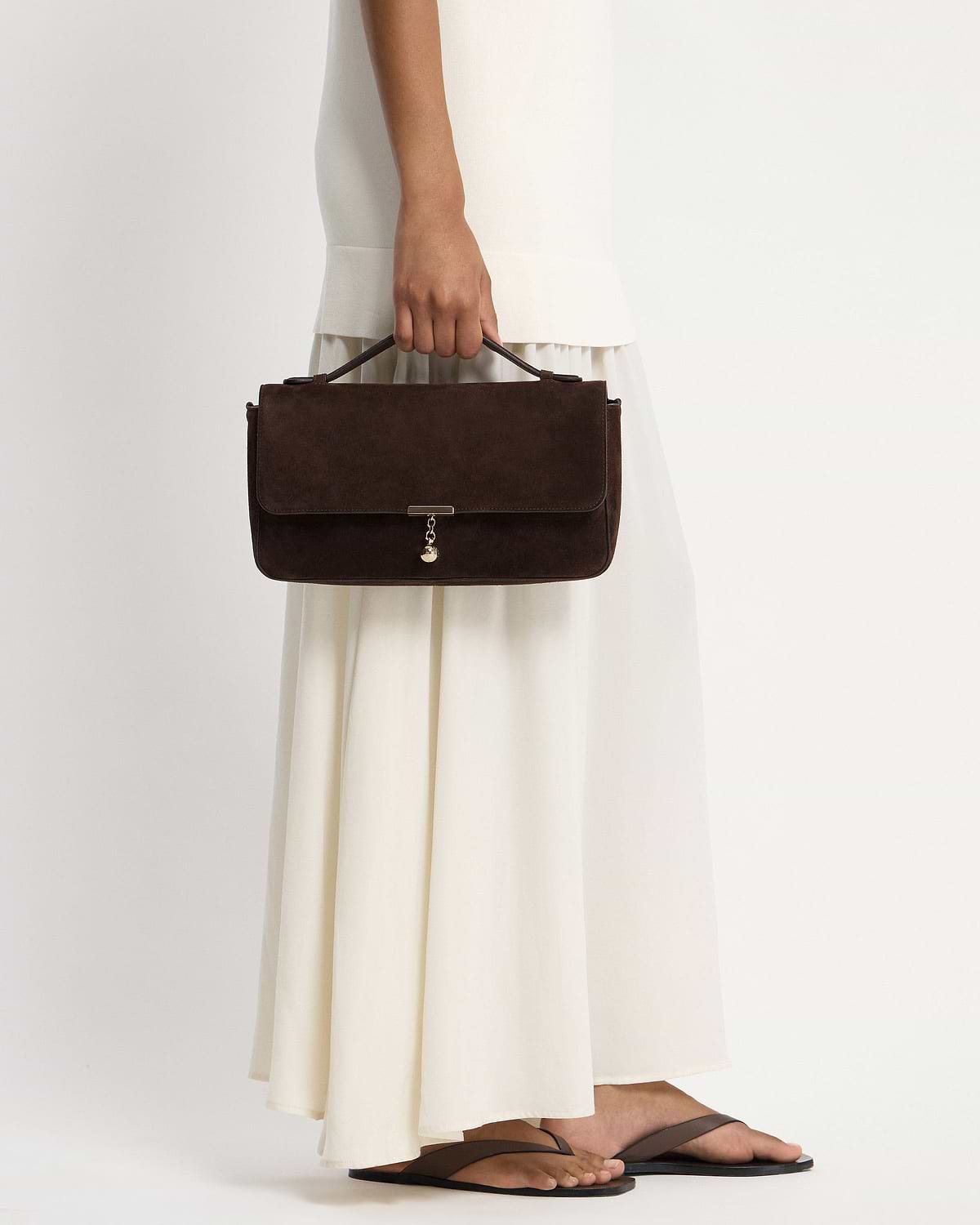 Hand holding espresso suede top-handle bag with gold charm, cream dress visible.