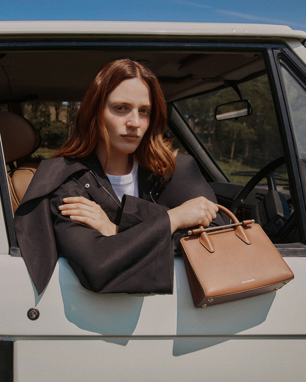 Woman in a car holding a chestnut Strathberry mini tote on the door.