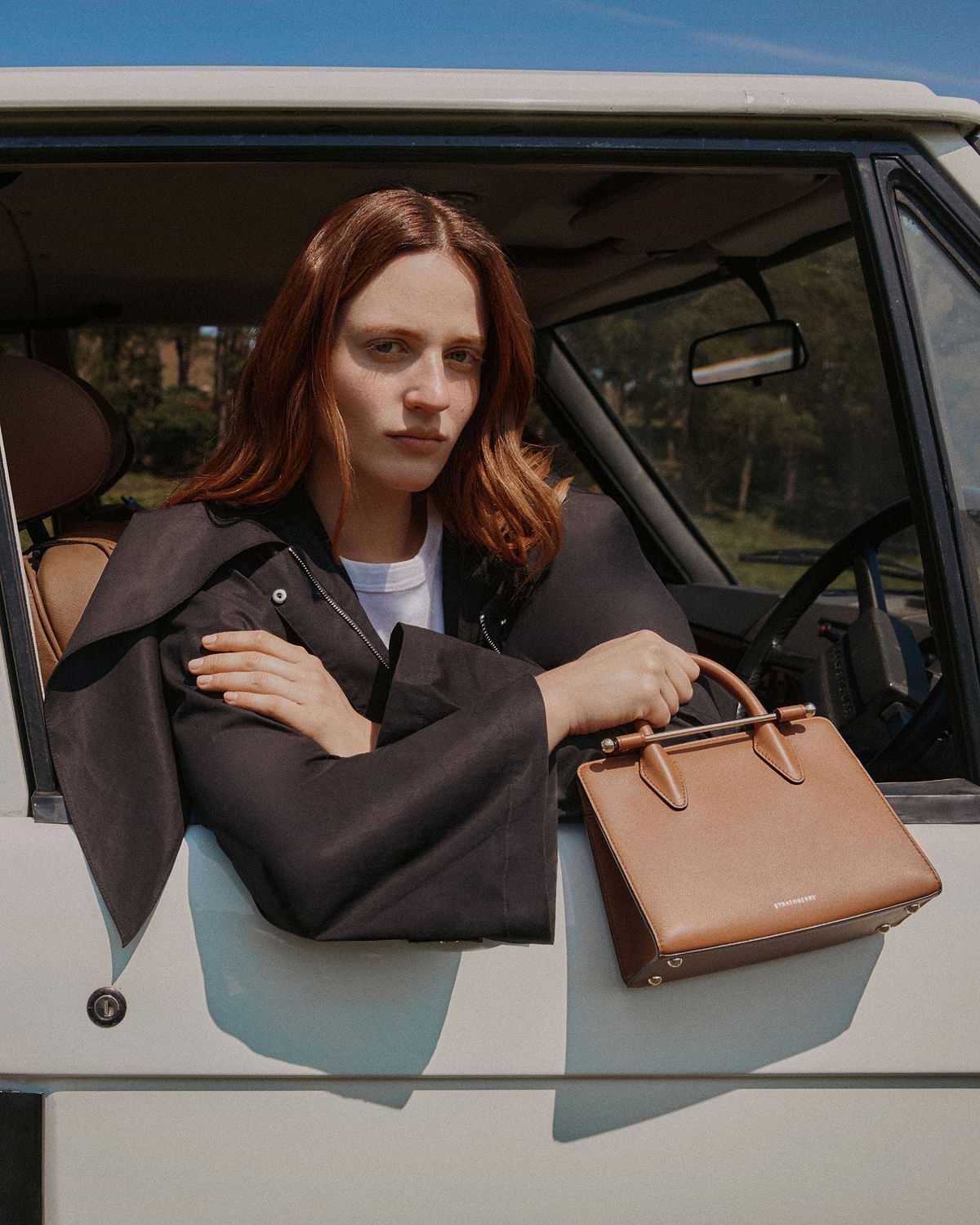 Woman in a car holding a chestnut Strathberry mini tote on the door.