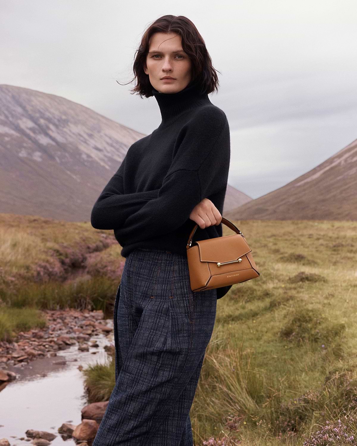 Woman in black jumper holding tan Strathberry Mosaic Nano bag with vanilla stitching.