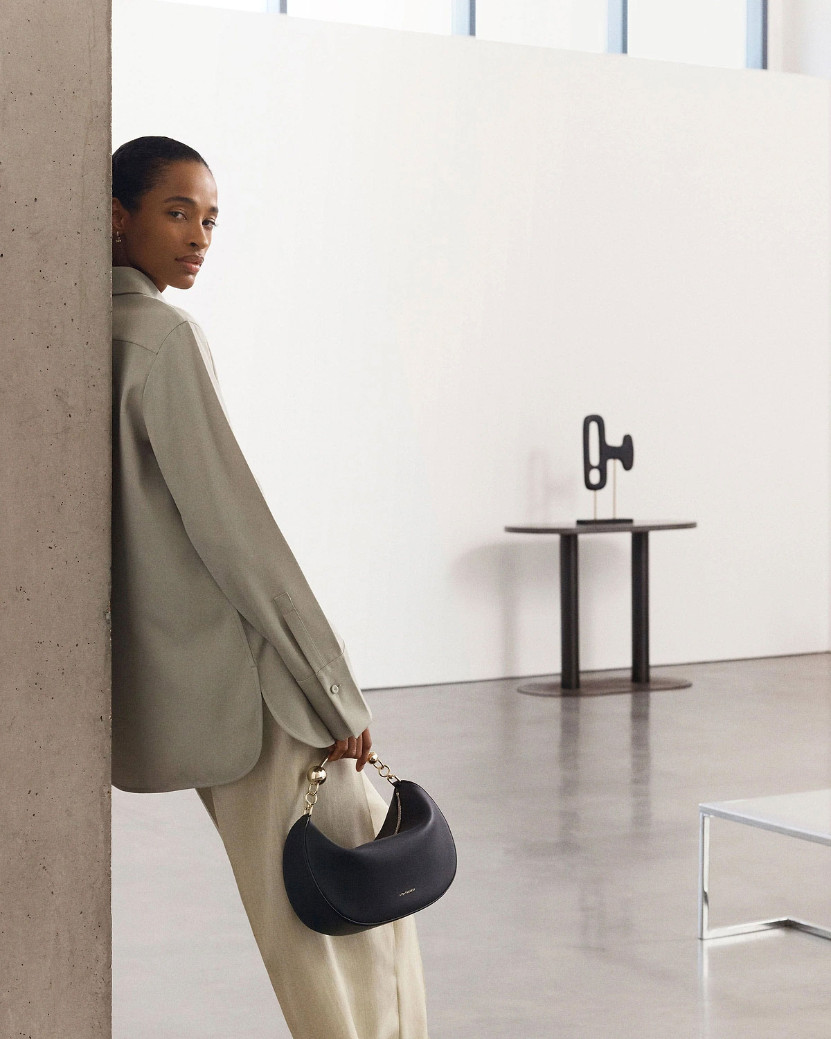 Woman leaning against a pillar holding a black crescent moon handbag with gold chain handle.