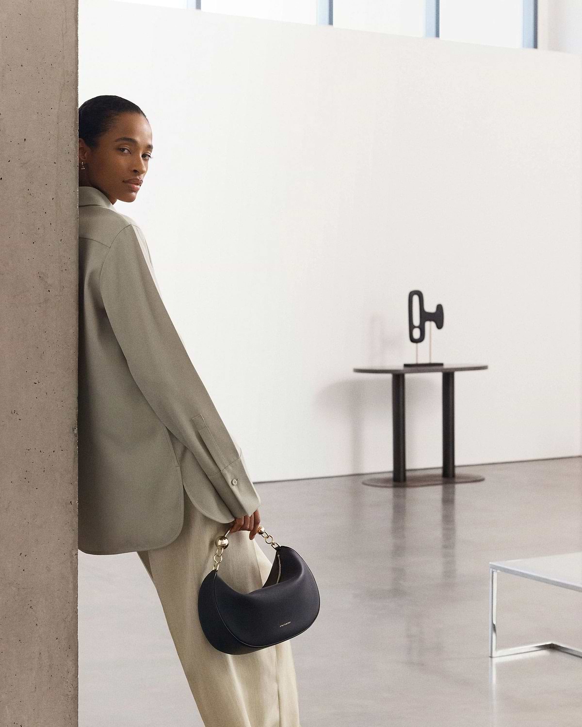 Woman leaning against a pillar holding a black crescent moon handbag with gold chain handle.
