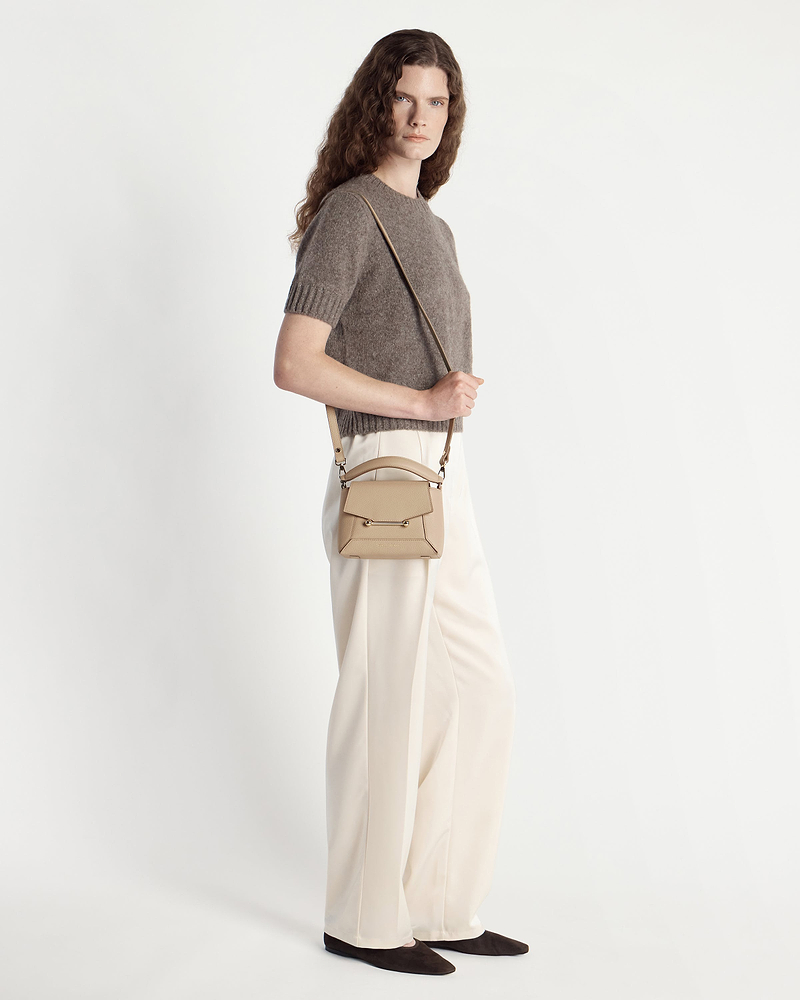 Model wearing a small sand-coloured structured top-handle crossbody bag with long strap over shoulder